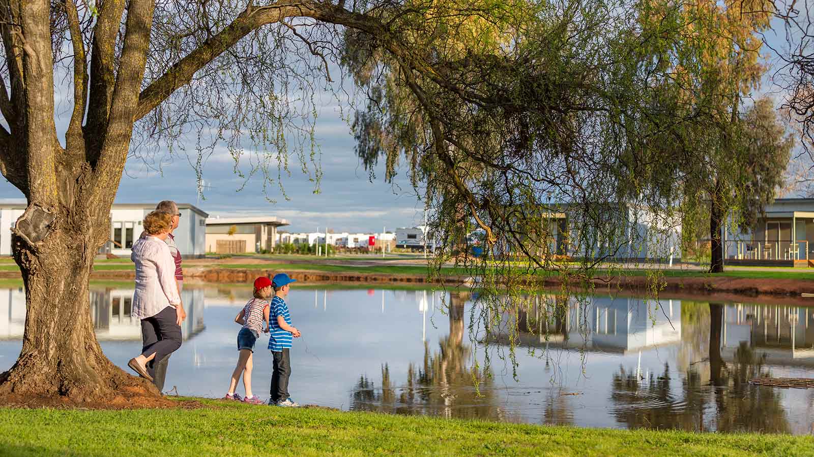 Parents and two children standing by a large tree at RACV Cobram Resort, while looking out at cabins across a lake.