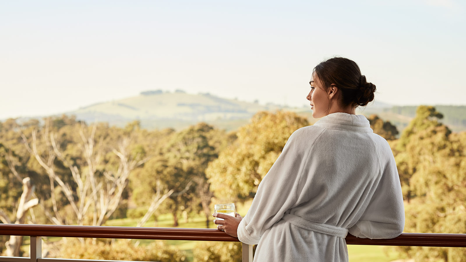 Person wearing a bathrobe and holding a glass while looking out from a balcony at RACV Goldfields Resort.