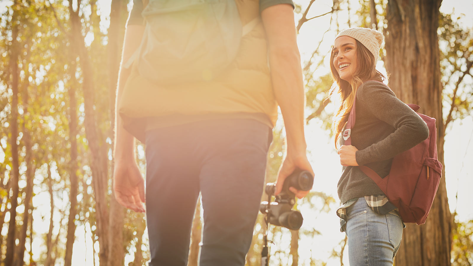 Two bushwalkers pause on a hiking trail near RACV Goldfields Resort, the woman on the right smiling back at her binocular-wielding companion on the left, with sunlight filtering through the tall native trees and the scene partially obscured by lens flare.