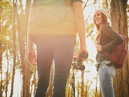 Two bushwalkers pause on a hiking trail near RACV Goldfields Resort, the woman on the right smiling back at her binocular-wielding companion on the left, with sunlight filtering through the tall native trees and the scene partially obscured by lens flare.
