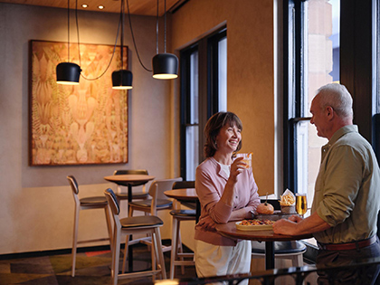 Couple smiling together by the window at RACV Hobart Hotel's Cascade on Collins bar, with pub style food on the table in front of them.