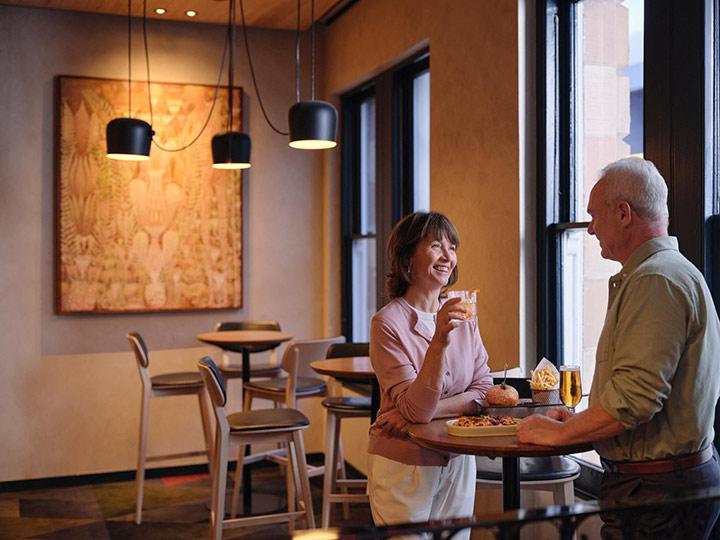 Couple smiling together by the window at RACV Hobart Hotel's Cascade on Collins bar, with pub style food on the table in front of them.