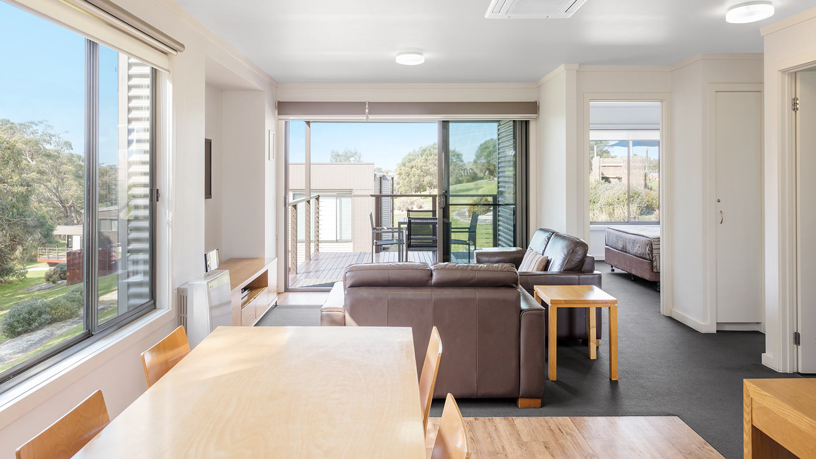 Lounge and dining area in a 2-bedroom cabin at RACV Inverloch Resort, featuring a balcony, television, couches and wooden dining table.