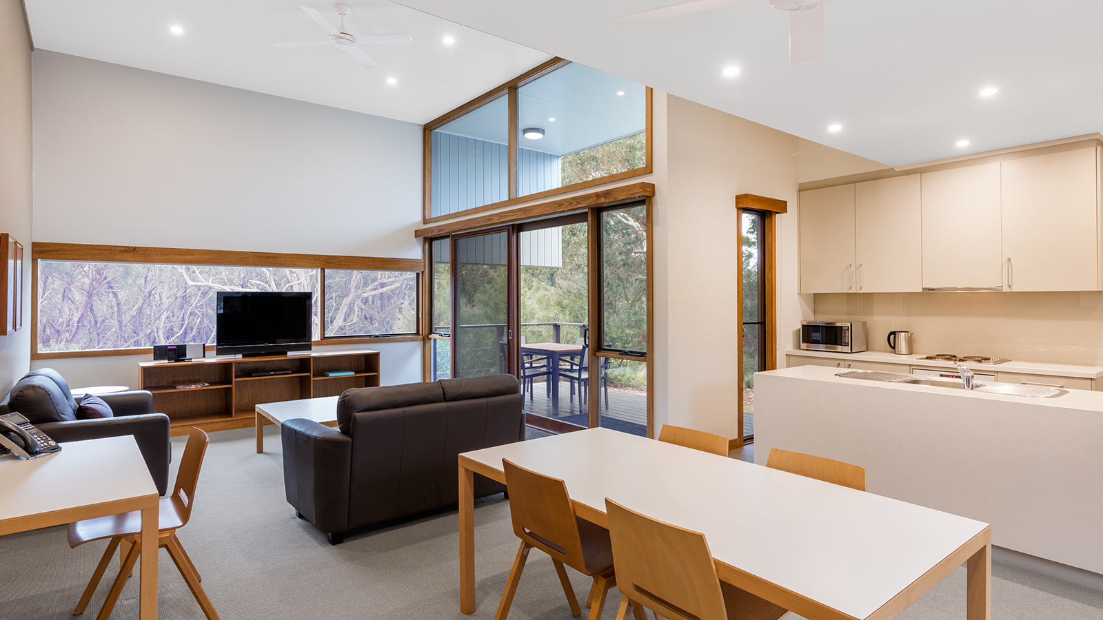 Kitchen and living area in a 2-bedroom villa at RACV Inverloch Resort, featuring a balcony, television and couches, wooden dining table and kitchen.