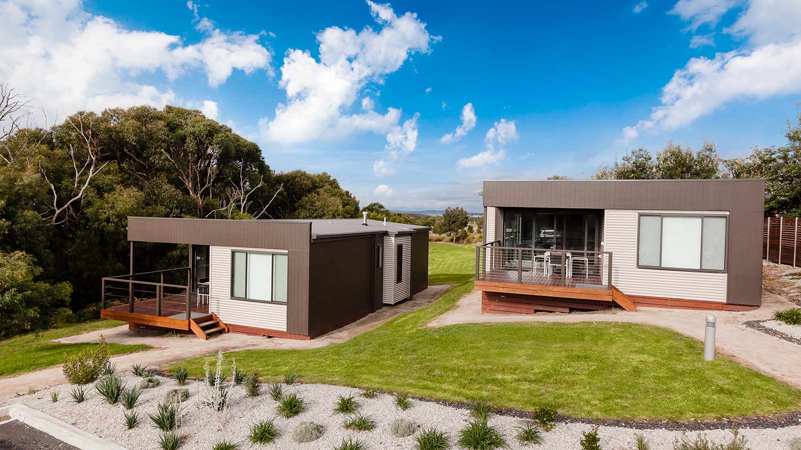 Two cabins set on a gradual hill at RACV Inverloch Resort, featuring wooden verandahs with tables and chairs.
