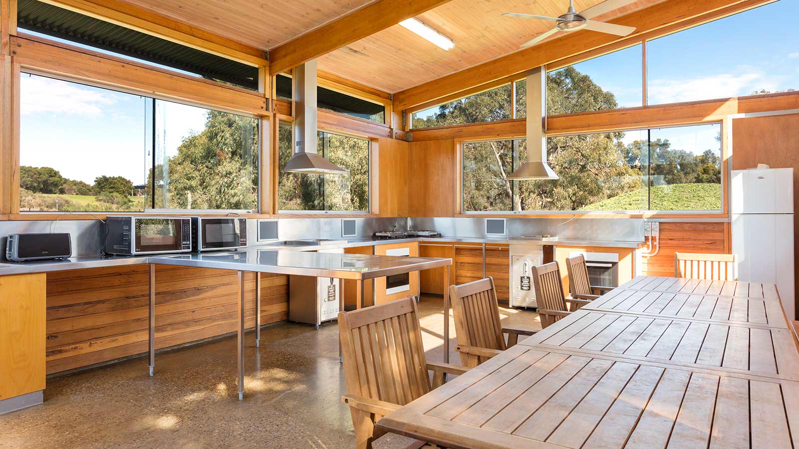 Shared kitchen facilities at RACV Inverloch Resort's caravan site, featuring a long wooden dining table, microwaves, toasters, ovens and gas cooktops.