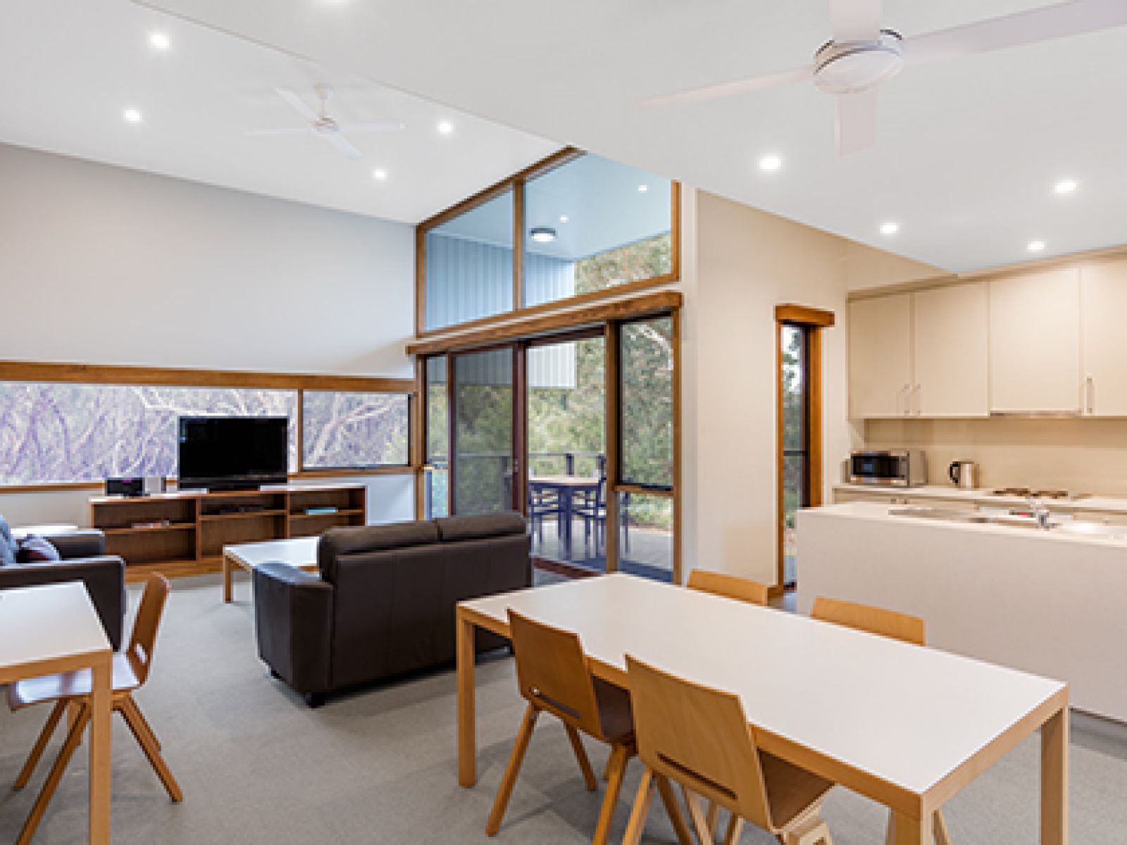 Kitchen and living area in a 2-bedroom villa at RACV Inverloch Resort, featuring a balcony, television and couches, wooden dining table and kitchen.