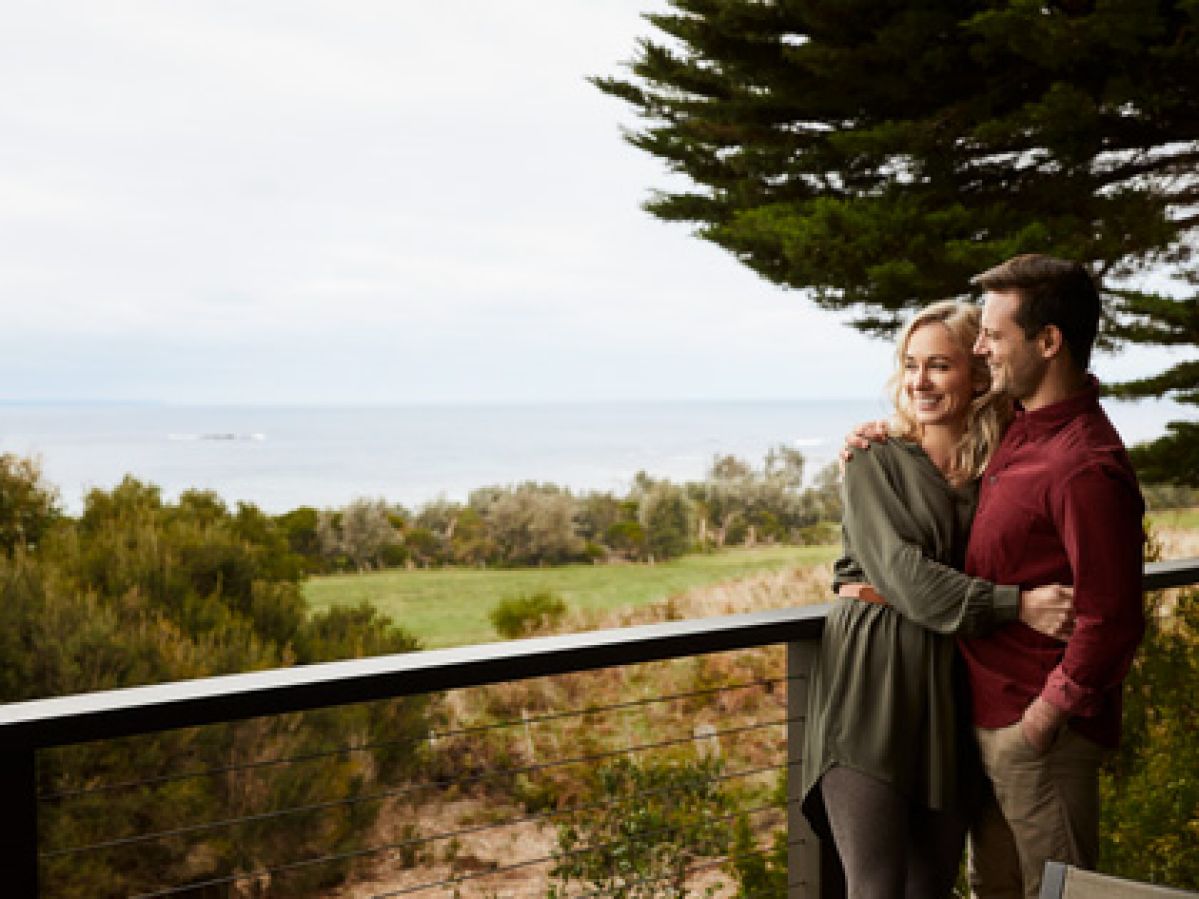 A couple hugging on a blacony overlooking grass and water. 