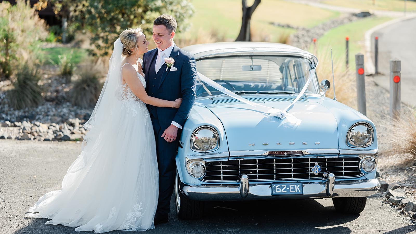 Bridge and groom posing in front of pale blue vintage Holden with wedding ribbon.