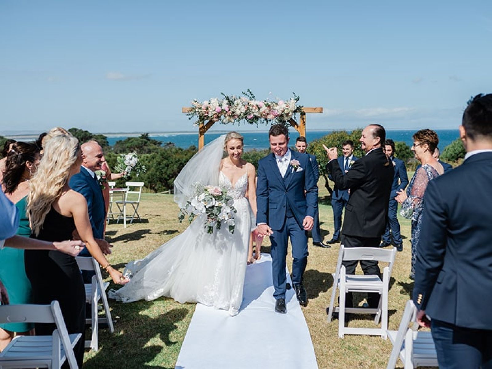 Outdoor wedding ceremony in front of sea at RACV Torquay resort, bride and groom walking down aisle as attendees applaud.