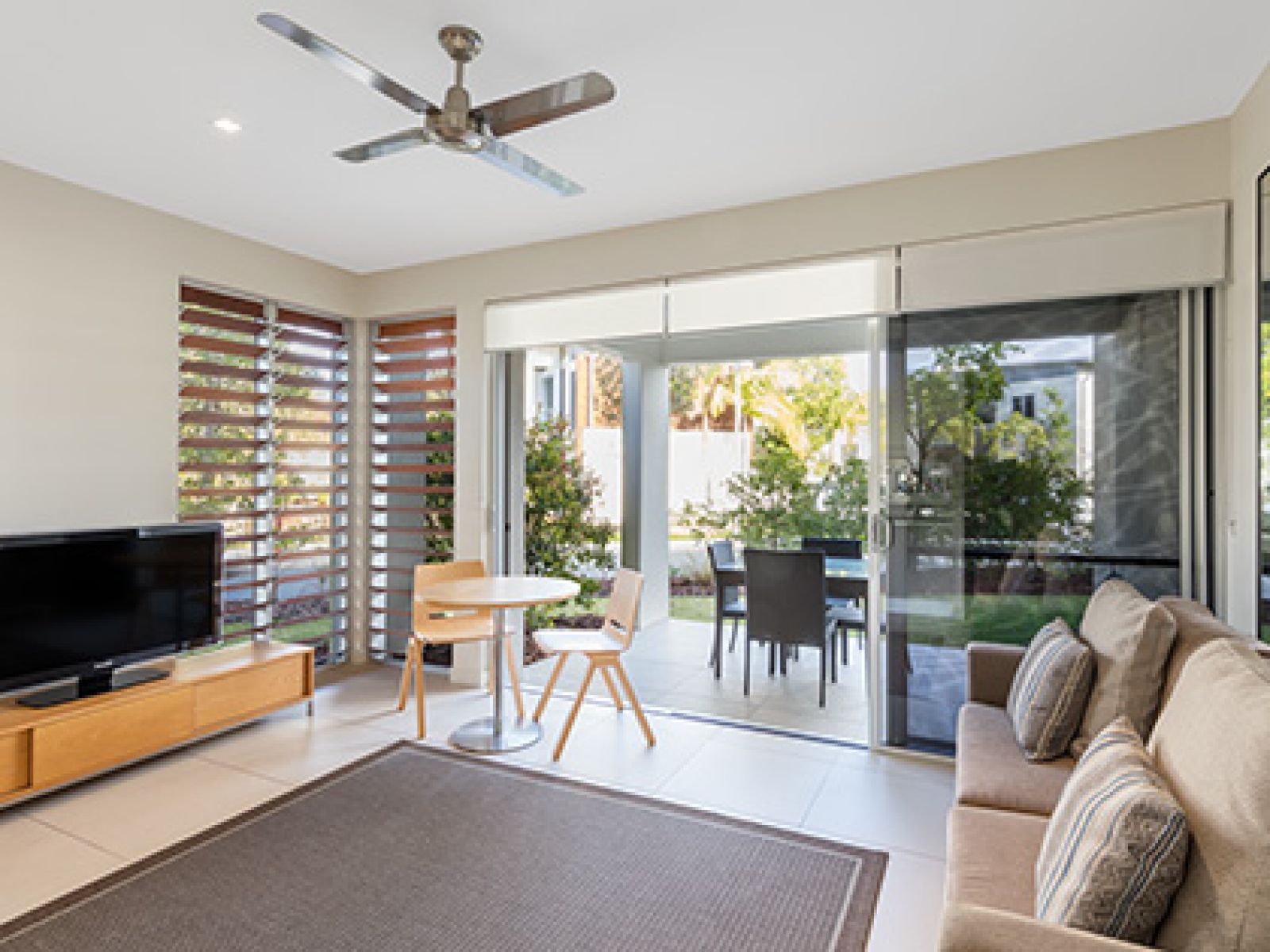 Noosa Resorts three bedroom sanctuary apartment living area, complete with a television, couch, table and chairs and sliding doors leading to the outdoor ground floor balcony. 