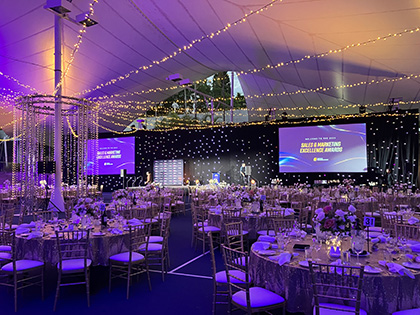 RACV Royal Pines Resort's tennis court transformed into a function space, with tables set up banquet style, a stage with screens and fairy lights hung up.