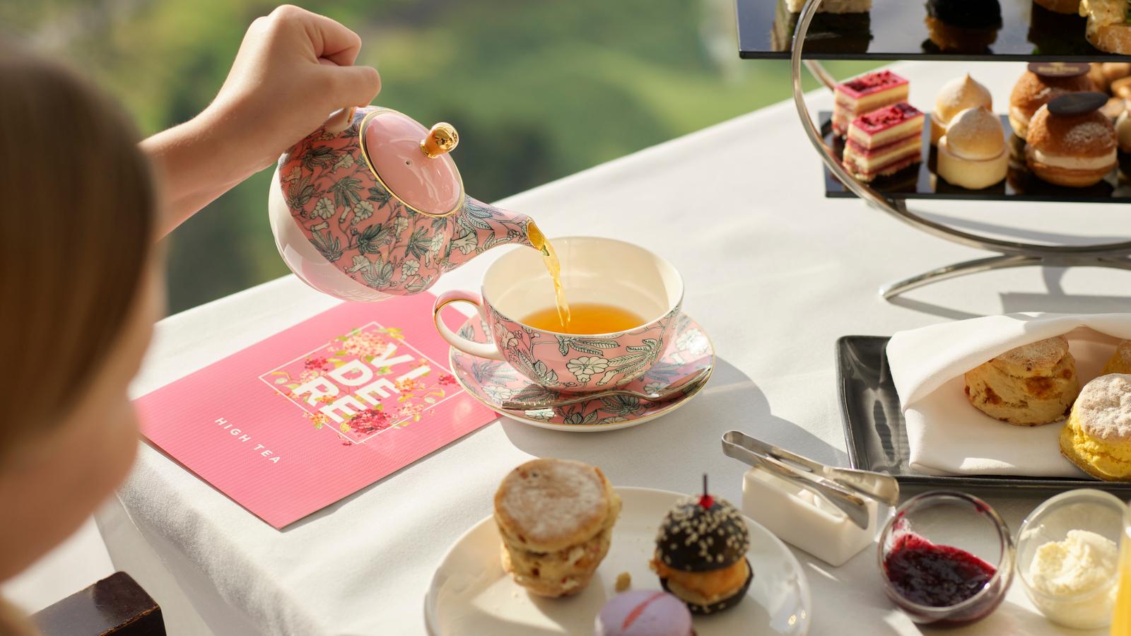 Girl pouring tea from a teapot on a table filled with small cakes.