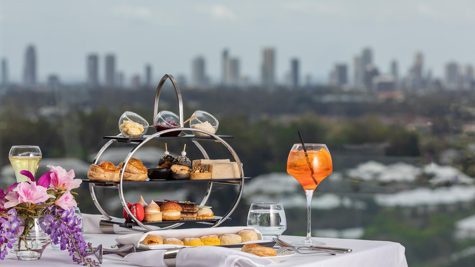 RACV Royal Pines Videre high tea setup with teapots, scones, and a tiered tray of desserts, savoury bites, jam, cream and butter, with a view of the city in the distance.