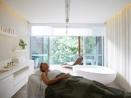 Person laying on their back with their eyes shut in a treatment room at RACV Royal Pines Resort's One Spa, next to a bathtub and outdoor patio.