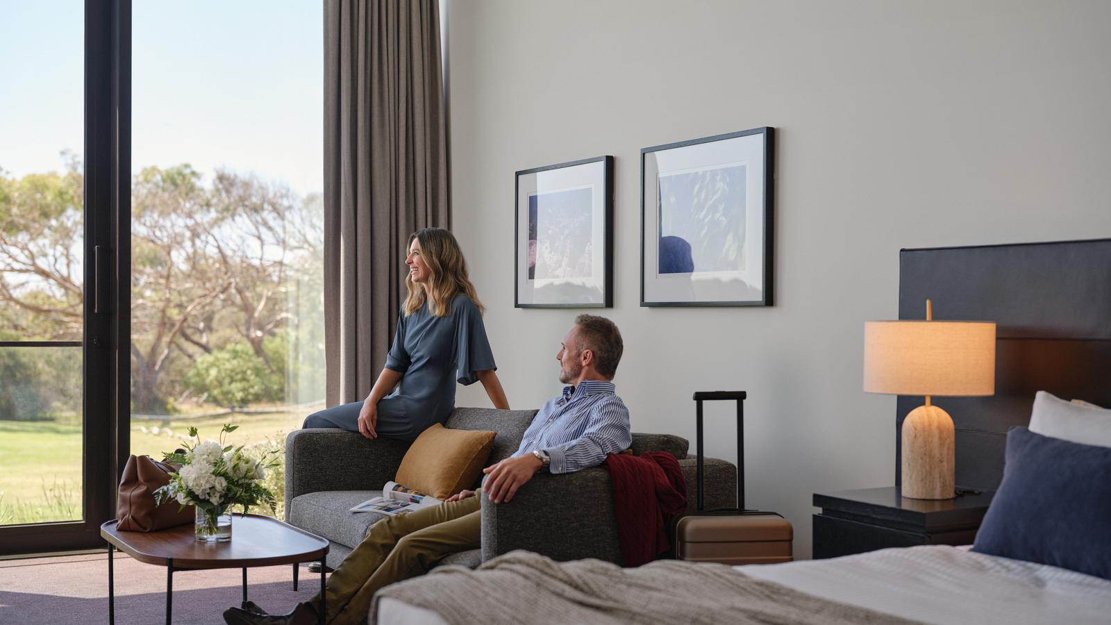 Couple relaxing in Golf View Room at RACV Torquay Resort, featuring a king bed, couches and desk.