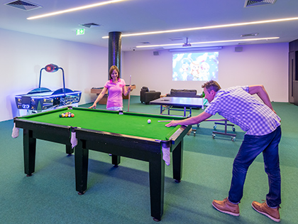 Two people playing pool at the RACV Torquay Resort games room, with a projector playing a movie on the wall behind them and table tennis, air hockey and foosball tables.