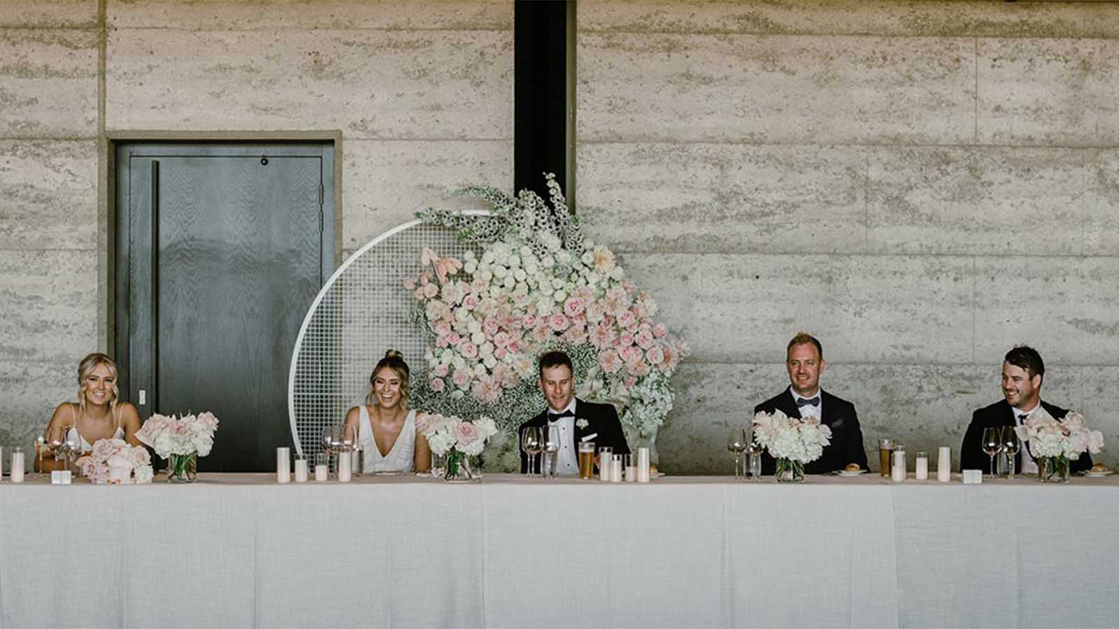Bridal party sitting on a a long table and smiling, in front of a flower arrangement.