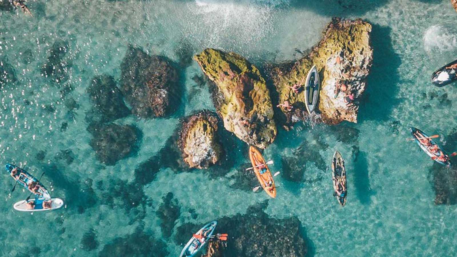 Aerial view of people kayaing in clear ocean waters near some rocks. 