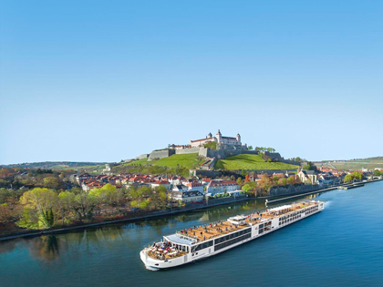 A crusie ship on a sunny day passing a small township with a castle. 