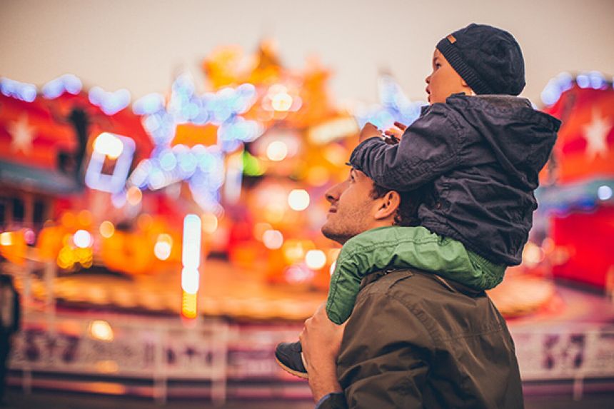 Child sitting on father's shoulders with amusement park lights in the background