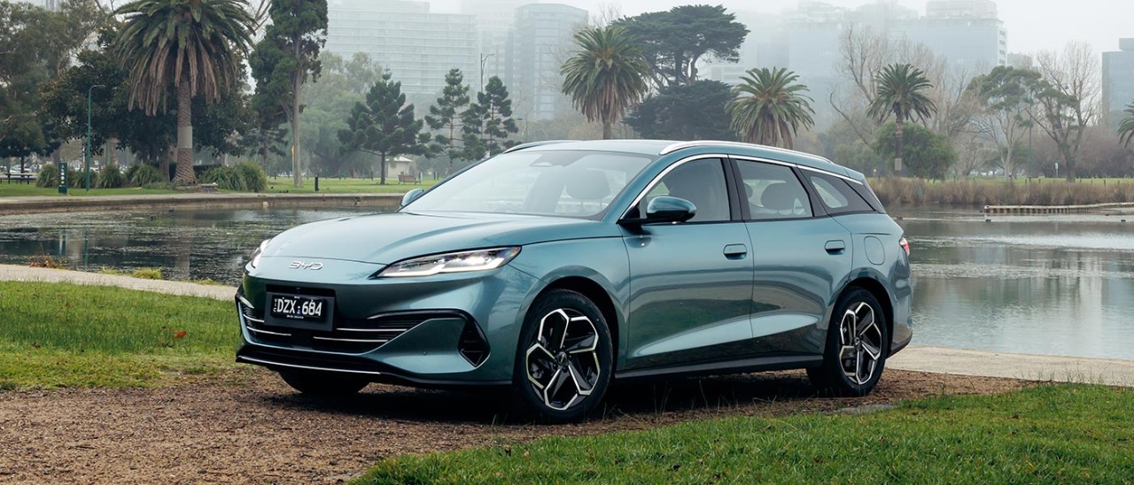 Sage green BYD Seal 6 Touring wagon parked at Albert Park Lake with misty Melbourne CBD skyline behind.