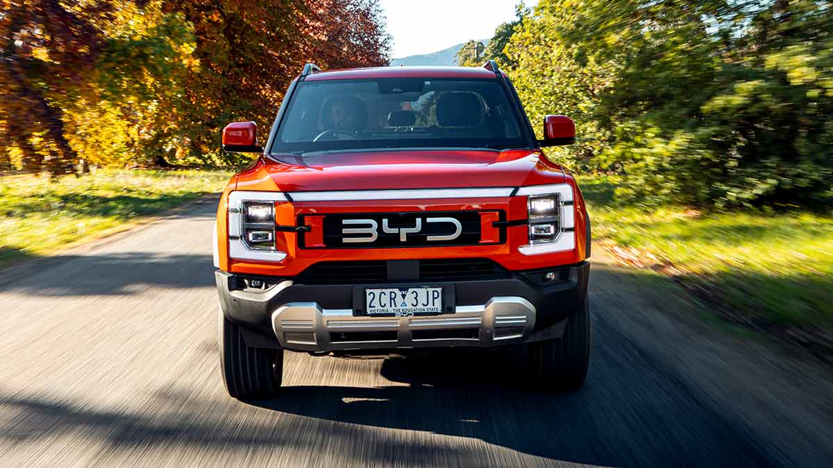 Front-on shot of orange BYD Shark 6 Performance ute driving on colourful autumn tree-lined road.