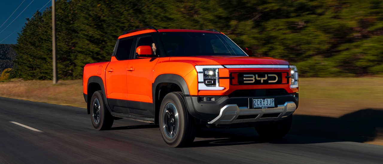 Orange BYD Shark 6 Performance ute driving on sealed country road with power lines and row of cypress trees behind.