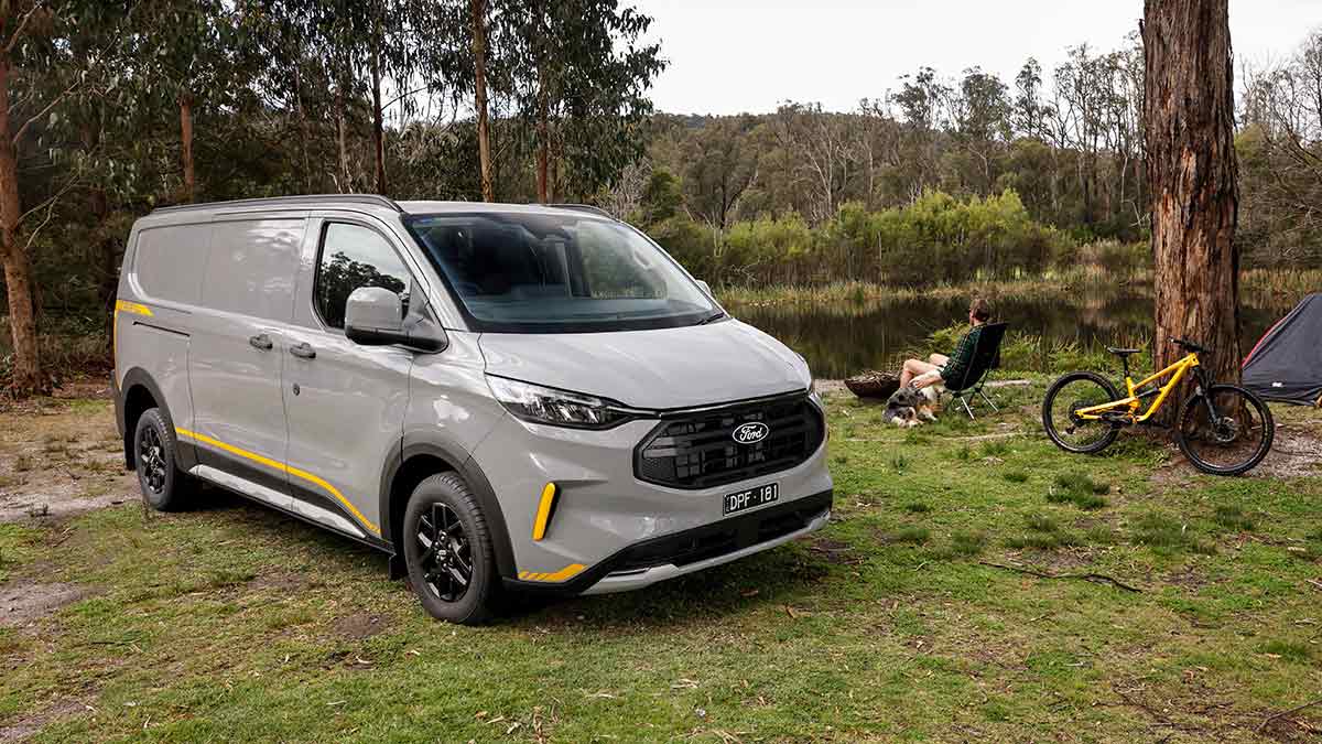 Silver Ford Transit Custom van at riverside campsite with man on deckchair next to dog and mountain bike.