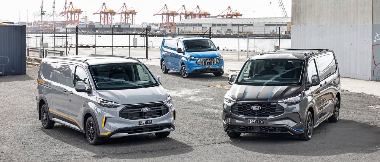Grey, silver and blue Ford Transit Custom Trail, EV and PHEV vans on dock in front of container terminal and gantry cranes.