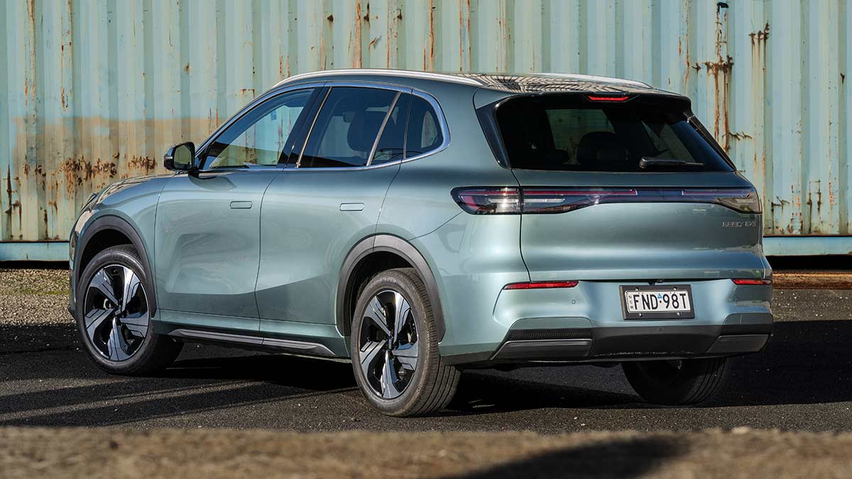 Rear three-quarter view of grey-blue Geely EX5 Inspire medium SUV in front of rusty shipping container