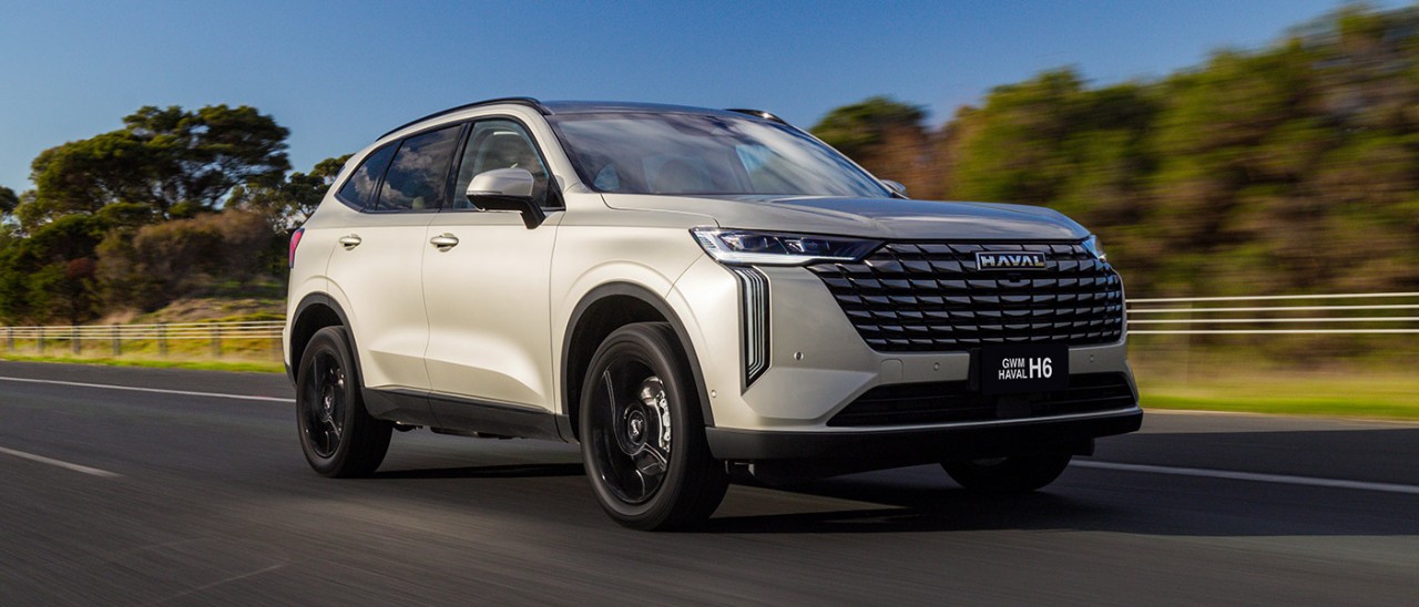 2025-GWM-Haval-H6-PHEV-banner-1400x600 Pearl-coloured GWM Haval H6 plug-in hybrid SUV on dual-lane highway in country Victoria with trees and blue sky behind.