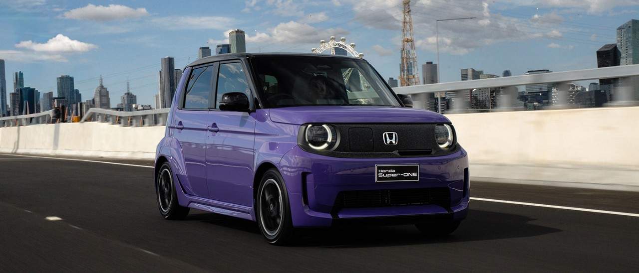 Purple Honda Super-One small boxy EV hatch on arterial road with city skyline in background.