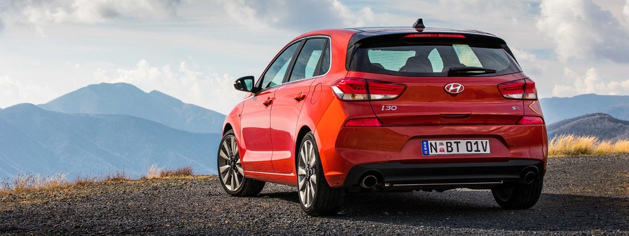Rear side view of a red Hyundai i30 SR Premium car parked on a gravel road with a mountain in the background