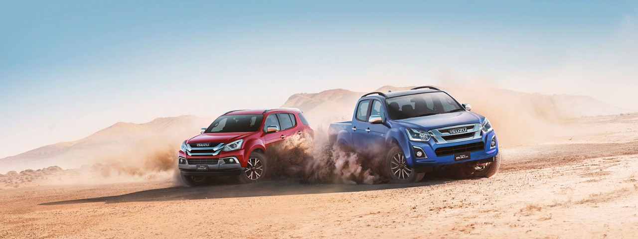 A red Isuzu MU-X next to a blue D-Max surrounded by dust clouds in a desert landscape