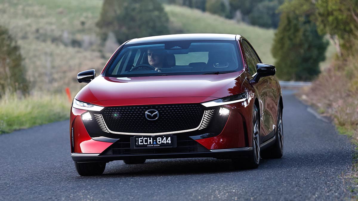 Red Mazda 6e EV sedan on unlined bitumen road in hilly farming district in country Victoria.