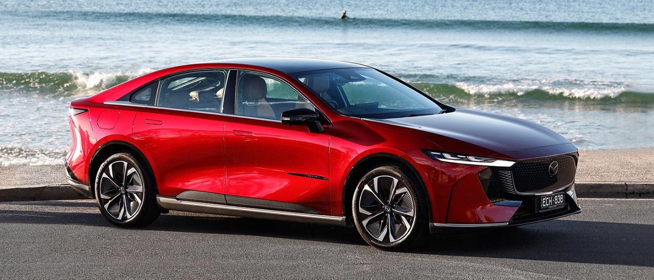 Red Mazda 6e electric sedan at beachfront carpark with lone surfer riding over wave in background.