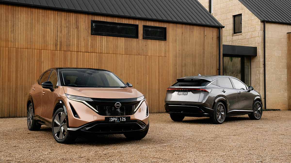 Copper-coloured Nissan Ariya Evolve and grey Nissan Ariya Advance+ SUVs in front of contemporary country house.