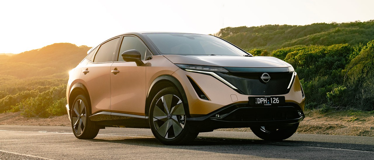 Copper-coloured Nissan Ariya electric SUV parked on side of coastal road with sun lighting up rear of car.