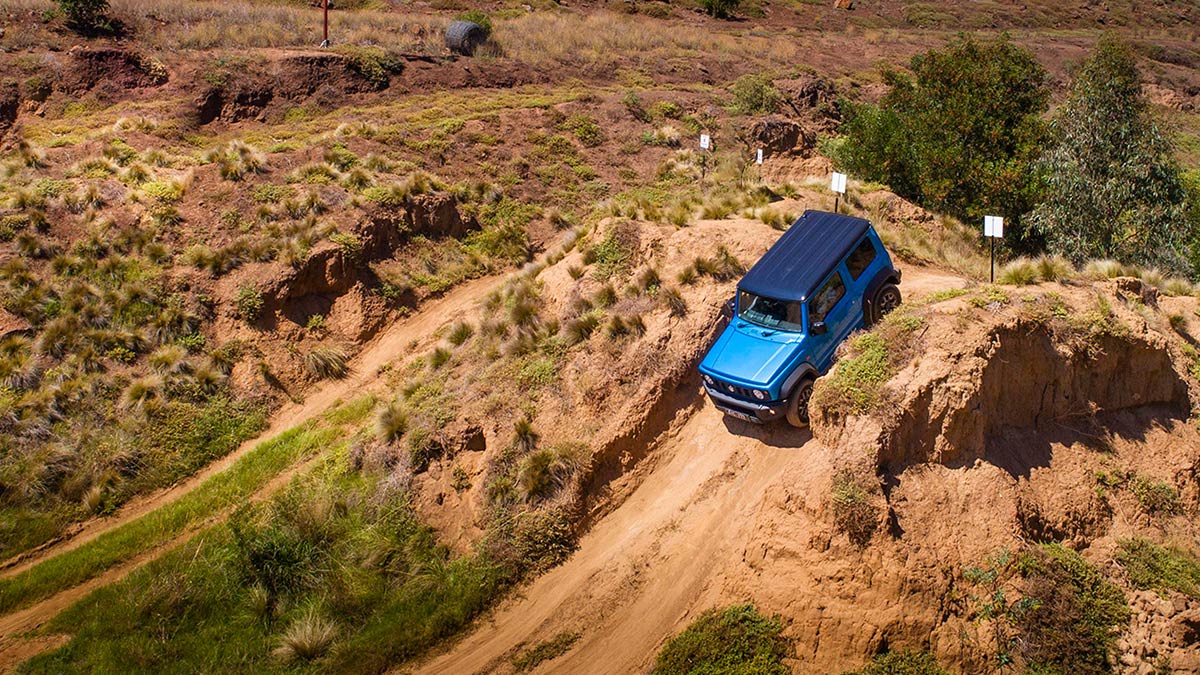 Distant top view of a blue Suzuki Jimny 2019 driving down a sandy hill dotted with shrubs