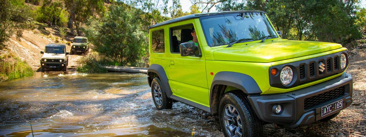 A lime green Suzuki Jimny 2019 car driving through a shallow body of water followed by two cars