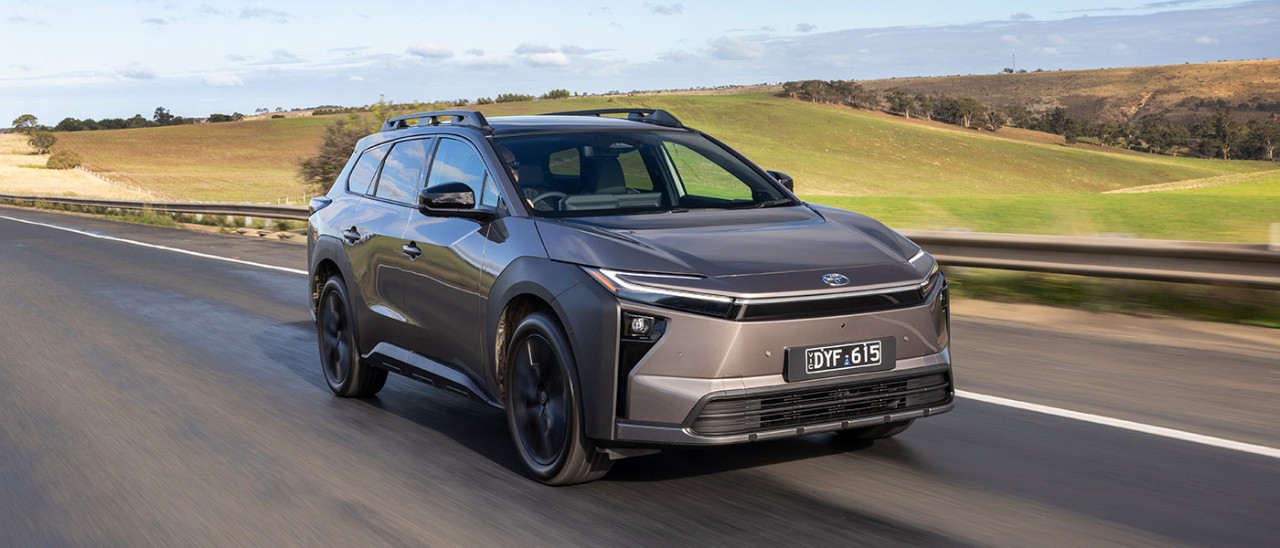 Bronze Toyota bZ4X Touring SUV driving on country highway next to undulating green paddocks and hillside.
