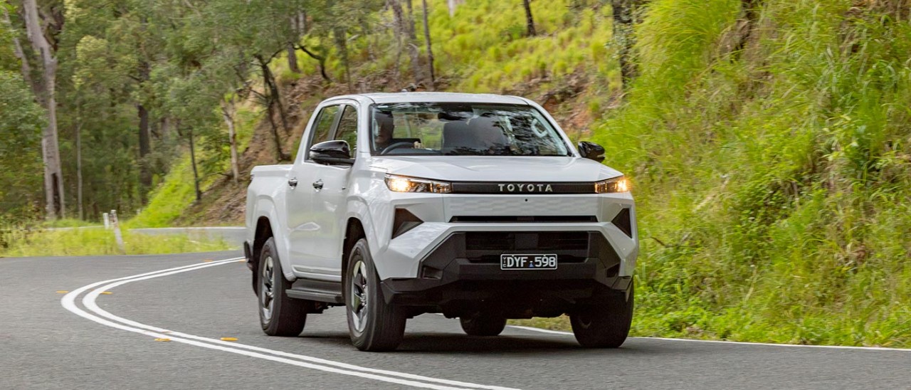 White Toyota HiLux electric dual-cab ute driving on winding high-banked country bitumen road.
