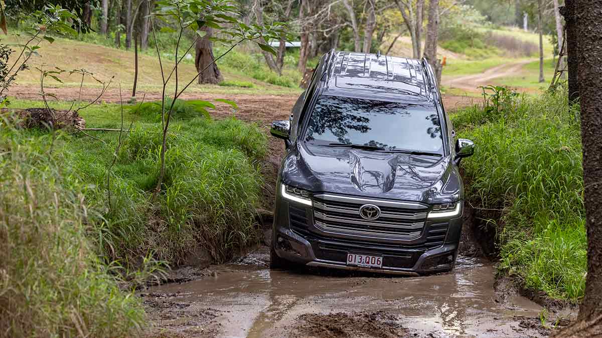 Dark grey metallic Toyota LandCruiser Sahara ZX Hybrid SUV descending creek bank at shallow muddy water crossing.