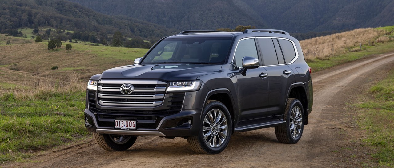 Dark grey metallic Toyota LandCruiser Sahara ZX Hybrid SUV on dirt road in alpine country.