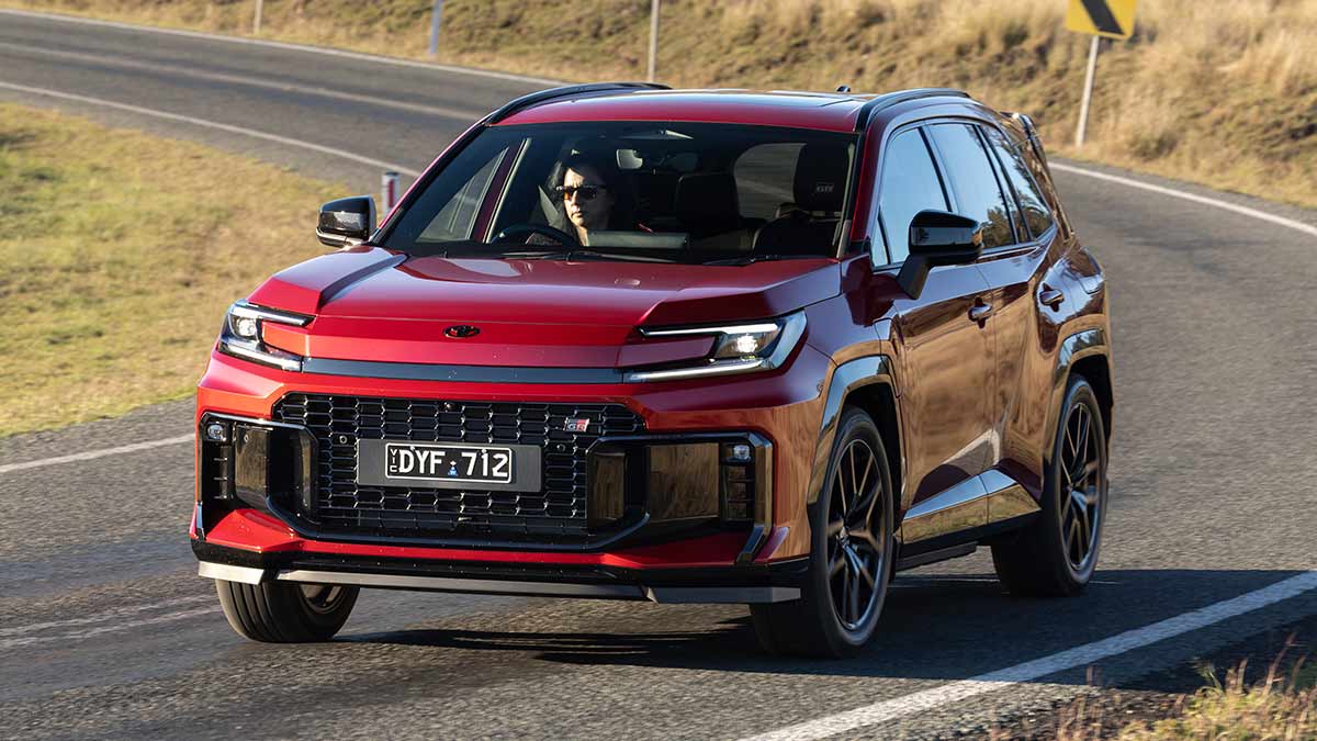 Red Toyota RAV4 GR Sport PHEV medium SUV going around corner on sunlit bitumen road with high grassy bank.