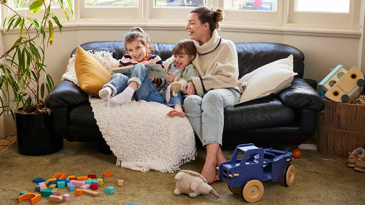 Mother and two young daughters having fun on couch in living room with toys strewn on floor