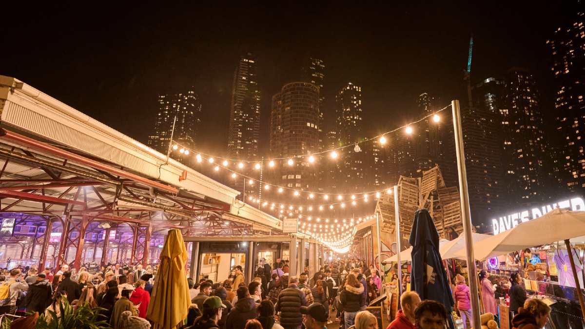 Throngs of people enjoying the free Winter Night Market at Queen Victoria market. Fairy lights and neon signs light up the market sheds while lights from Melbourne's city skyscrapers can be seen in the background