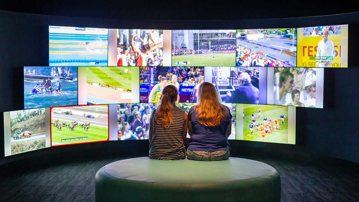 Two people looking at multiple screens showing sporting events at the Melbourne Sports Museum in Melbourne.