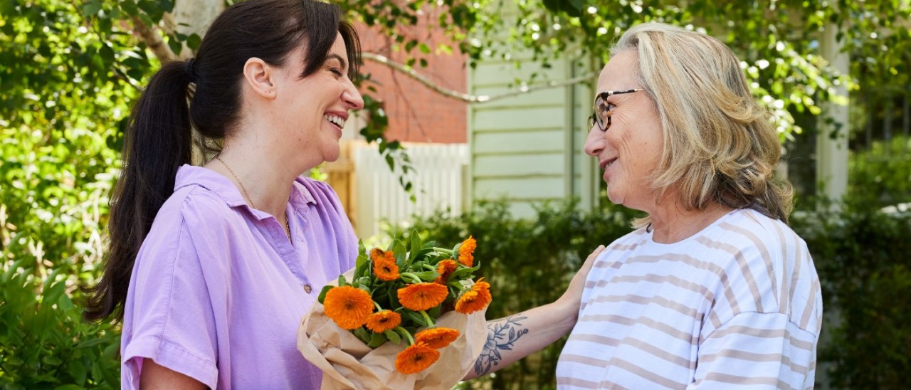 Two women, one young with long dark hair in a ponytail and one middle-age with greying blonde hair stand outside under a tree. The younger woman has brought bright orange flowers as a gift and both women are smiling